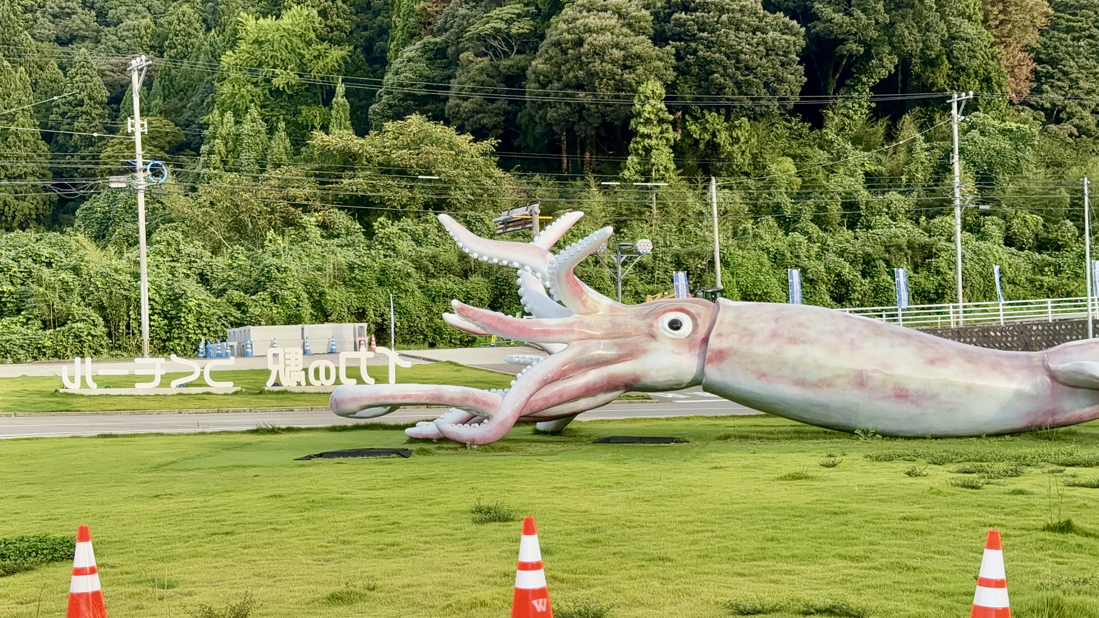 芝生の上に寝そべる巨大なイカのモニュメント。触腕を大きく広げ、奥に「イカの駅つくモール」の文字看板