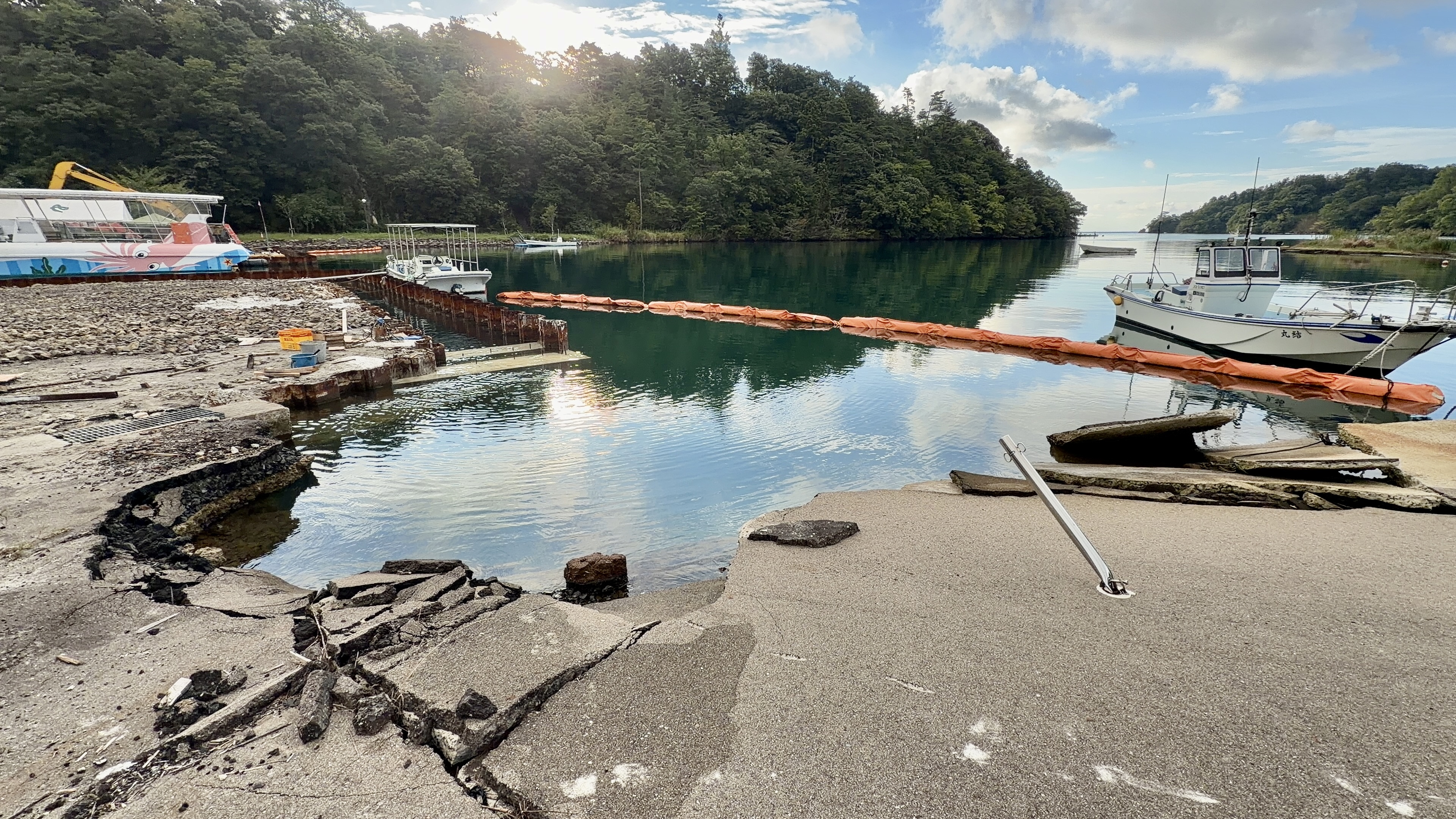 地震で割れてひび割れた護岸のコンクリート、水がたまり冠水している様子。奥にオレンジのオイルフェンスと漁港