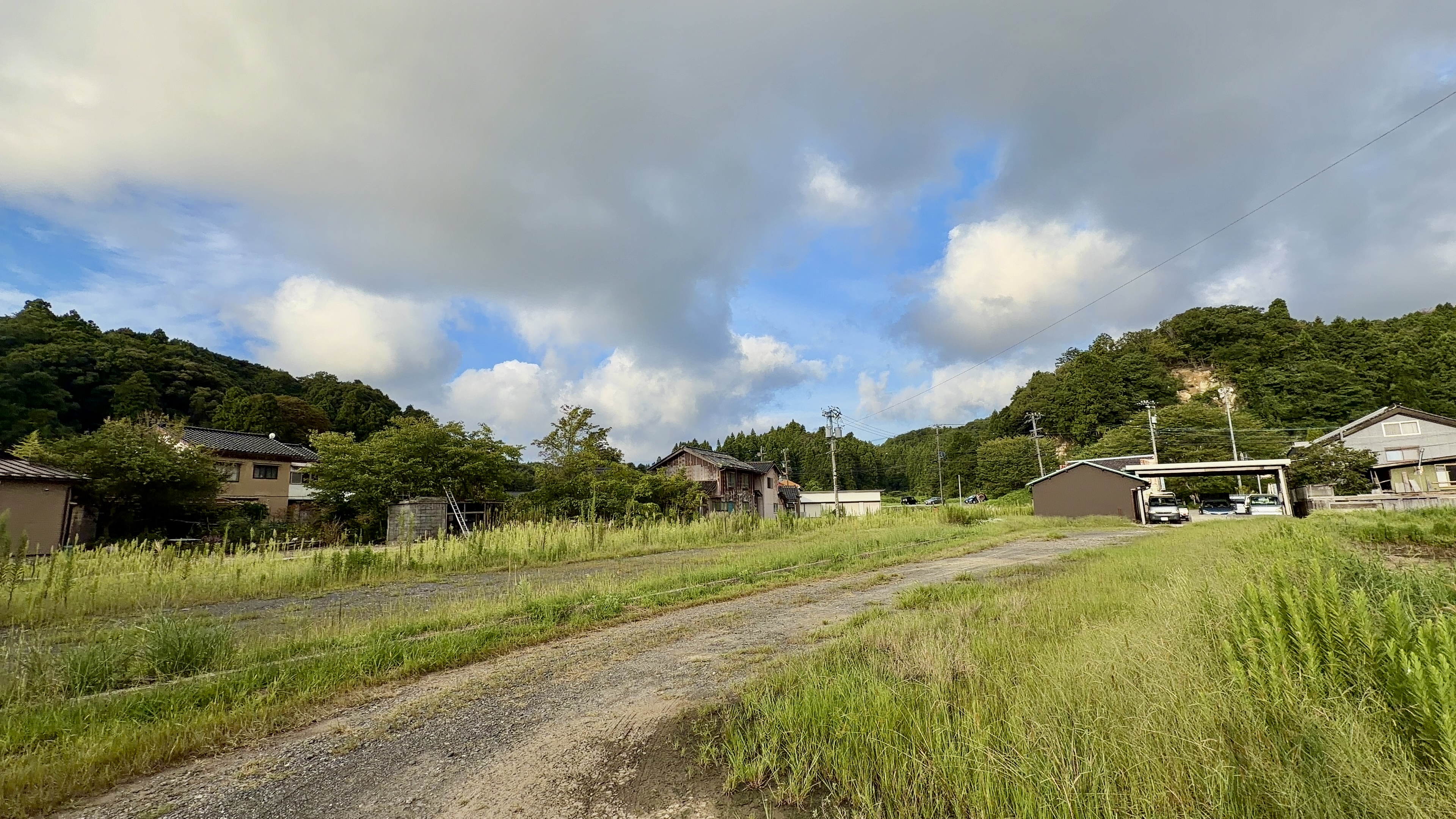 越坂の集落を抜ける未舗装の道、両脇に民家と草むら、奥に山並み