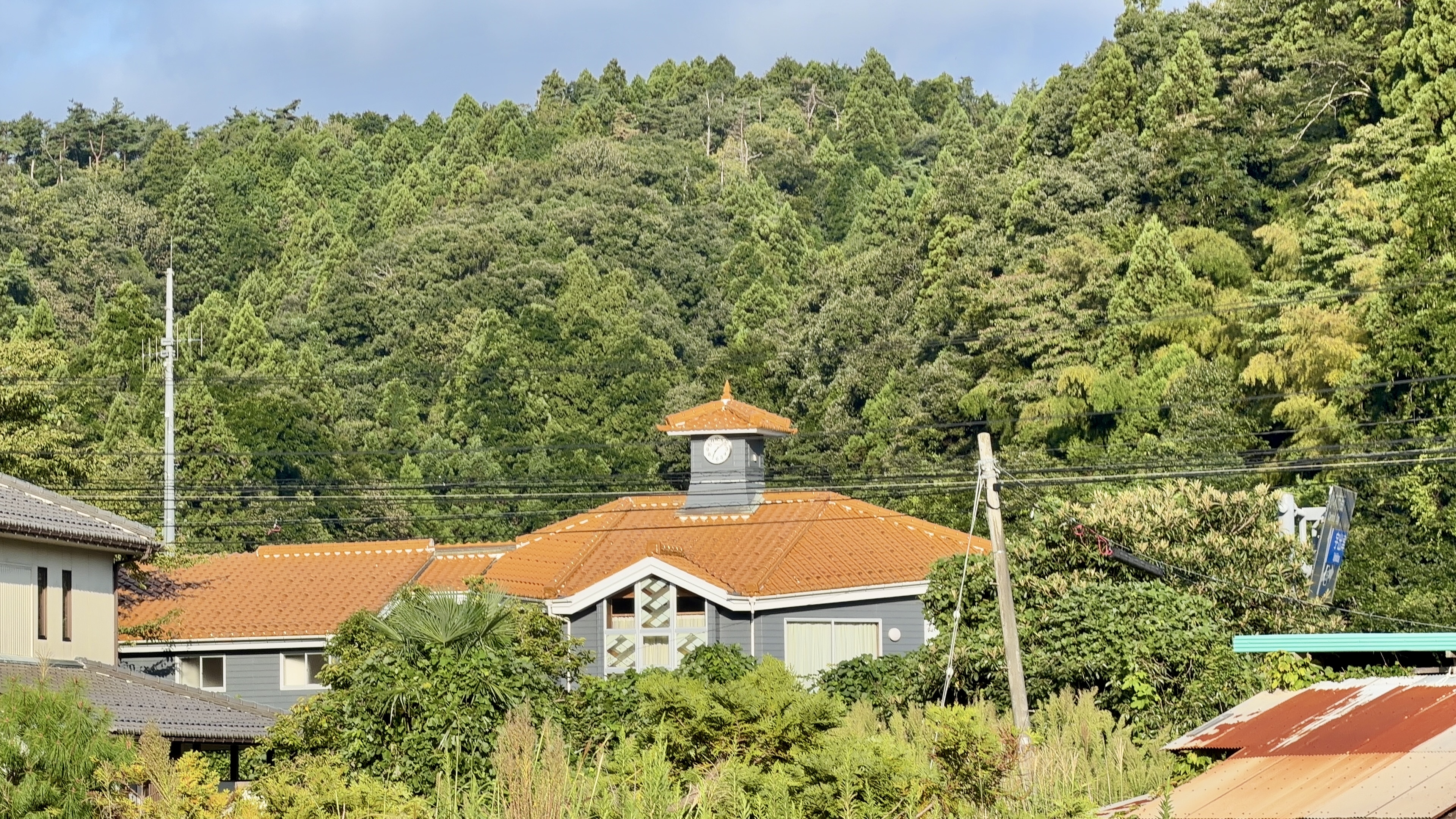 オレンジ色の瓦屋根に小さな時計塔がのった建物、周りを深い緑の山に囲まれている